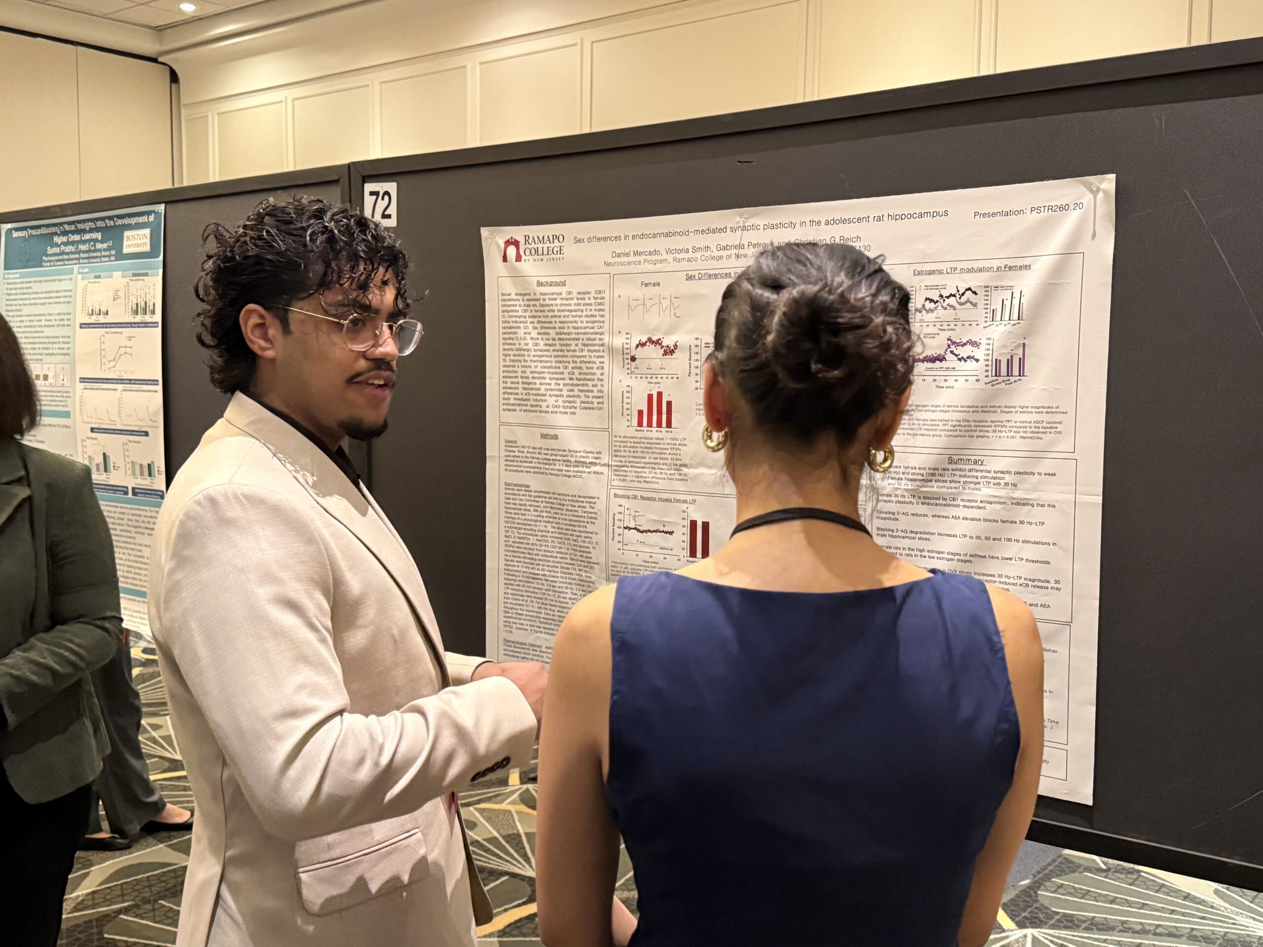 Two people stand in front of a scientific poster and discuss its content at a conference. The man on the left gestures while talking; the woman on the right has her back to the camera. Other posters are visible in the background.