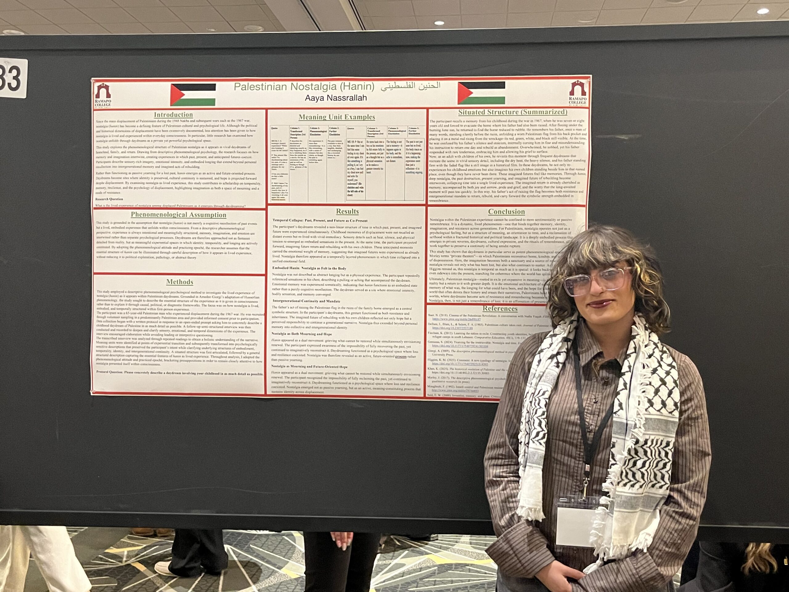 A woman stands and smiles next to a research poster titled “Palestinian Nostalgia (Hanin)” at a conference. The poster features text, Palestinian flags, and red and green borders. The woman wears glasses and a keffiyeh scarf.