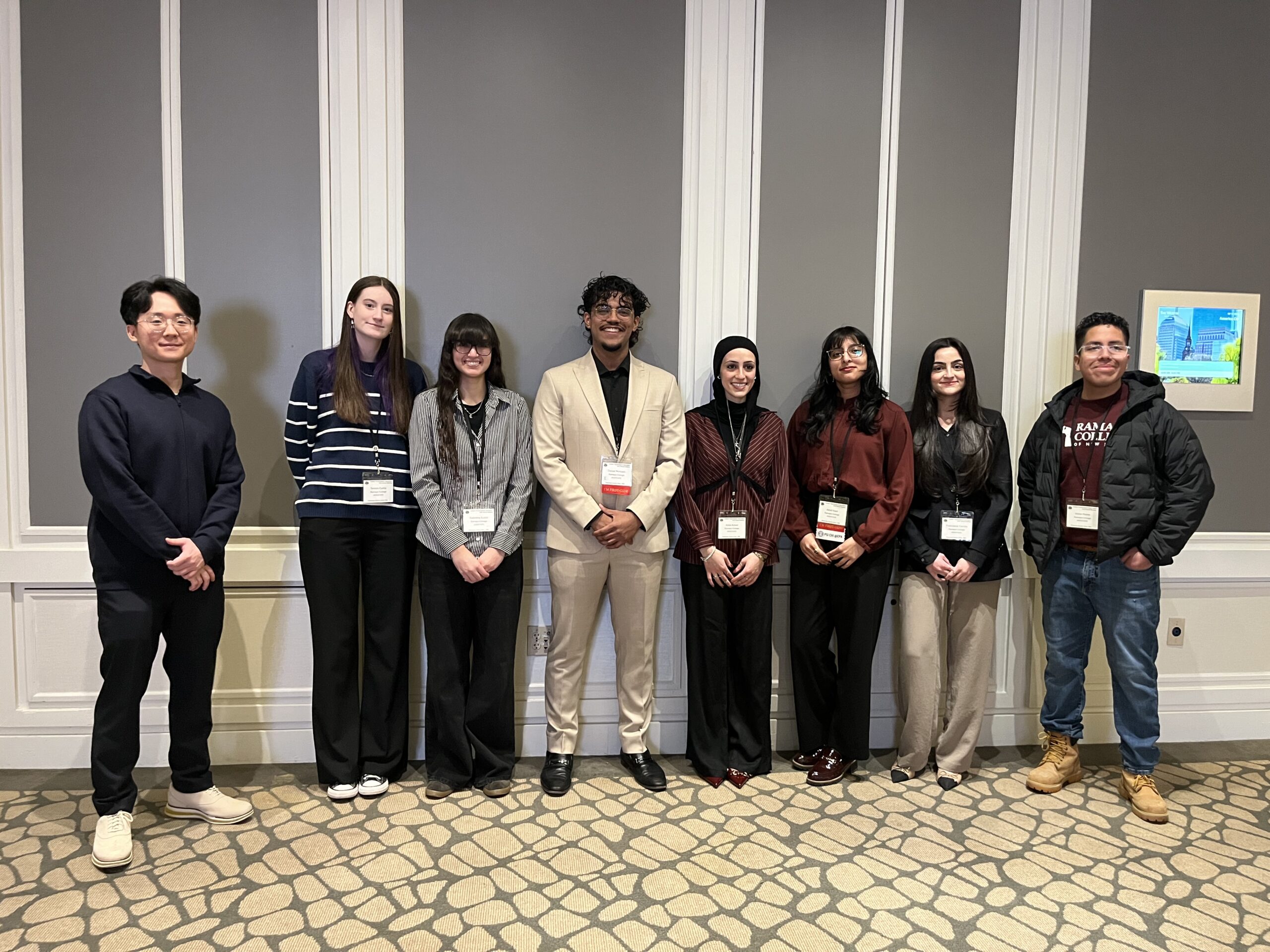 Eight people stand in a row indoors, smiling at the camera. They wear business or casual attire and conference badges. The background features white panel walls, a gray accent, and a framed picture on the right.