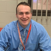 Stephen sits at a desk. He is wearing a blue button down shirt. He has a lanyard around his neck. He is smiling at us.