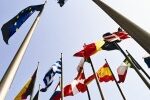 Multiple national and European Union flags on flagpoles wave against a bright, clear sky, viewed from below.