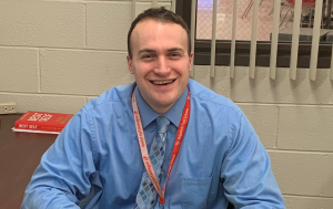 Stephen sits at a desk. He is wearing a blue button down shirt. He has a lanyard around his neck. He is smiling at us.