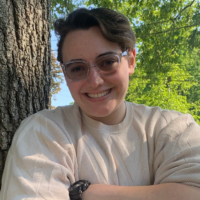 Lauren Marzano's headshot. She is leaning against a tree on her right side and has her arms folded across her chest.