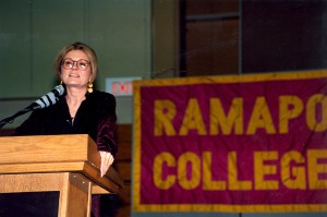 Gloria Steinem speaks at a podium in front of a Ramapo College banner in maroon and gold.