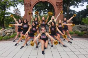 Ramapo College Dance Team in uniform with pom poms posing in front of The Arch.