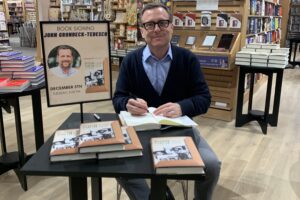 American Studies Professor John Gronbeck-Tedesco sits at a table signing a copy of his book inside Barnes & Noble bookstore.