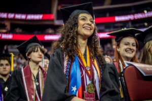 A smiling Stephanie Viera '25 is seated among graduates at the 2025 Commencement wearing a graduation robe, and several stoles, cords, and medals