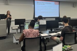A career advisor presents job search advice in front of students. The advisor is at the front of a classroom and the students are facing the front of the classroom with their backs to us.