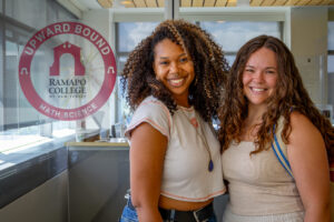 Two women who are graduate students stand next to each other in front of a sign for the Upward Bound office.