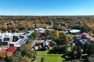 Aerial view of the Ramapo College campus. The sky is various shades of vibrant blue with trees bearing green needles and yellow, red, and orange leaves. A green grassy hill is in the foreground with the historic Birch Mansion to the right and the Academic Complex, topped by solar panels, to the left and center back.