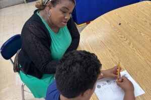 A woman wearing a teal dress and black cardigan sits at a table with a boy while he works on a worksheet.