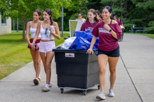 A group of four female college students walk down a path with a large bin on wheels filled with items for a dorm room.