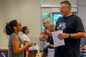A woman wearing a green vest looks up at and speaks to a tall man wearing a blue T-shirt, holding a piece of paper.
