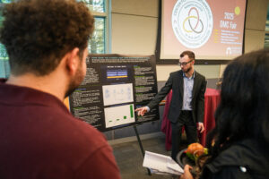 A student wearing a suit points to a research poster and speaks to guests about his research. Behind him is a projector screen displaying the DMC Center logo with the words 2025 Fair.